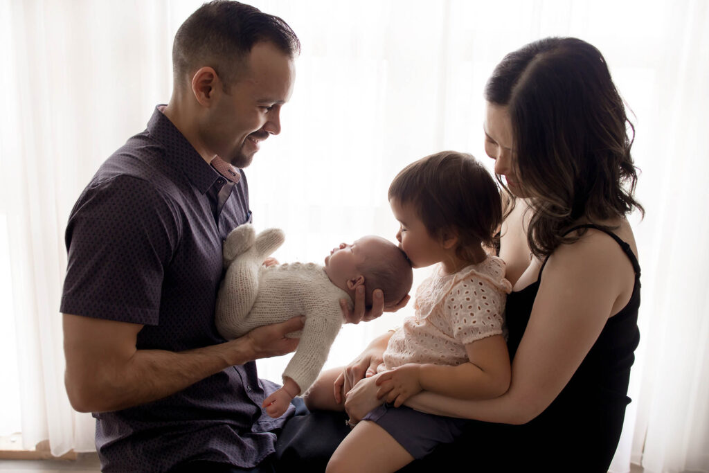 Family sitting by a bright window; toddler kisses baby while parents hold their children, facing each other.
