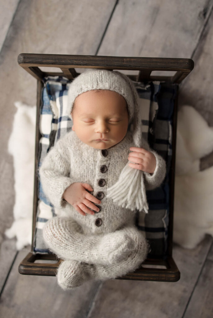 Sleeping newborn in white knitted outfit and hat on wooden bed with plaid blanket and cushion.