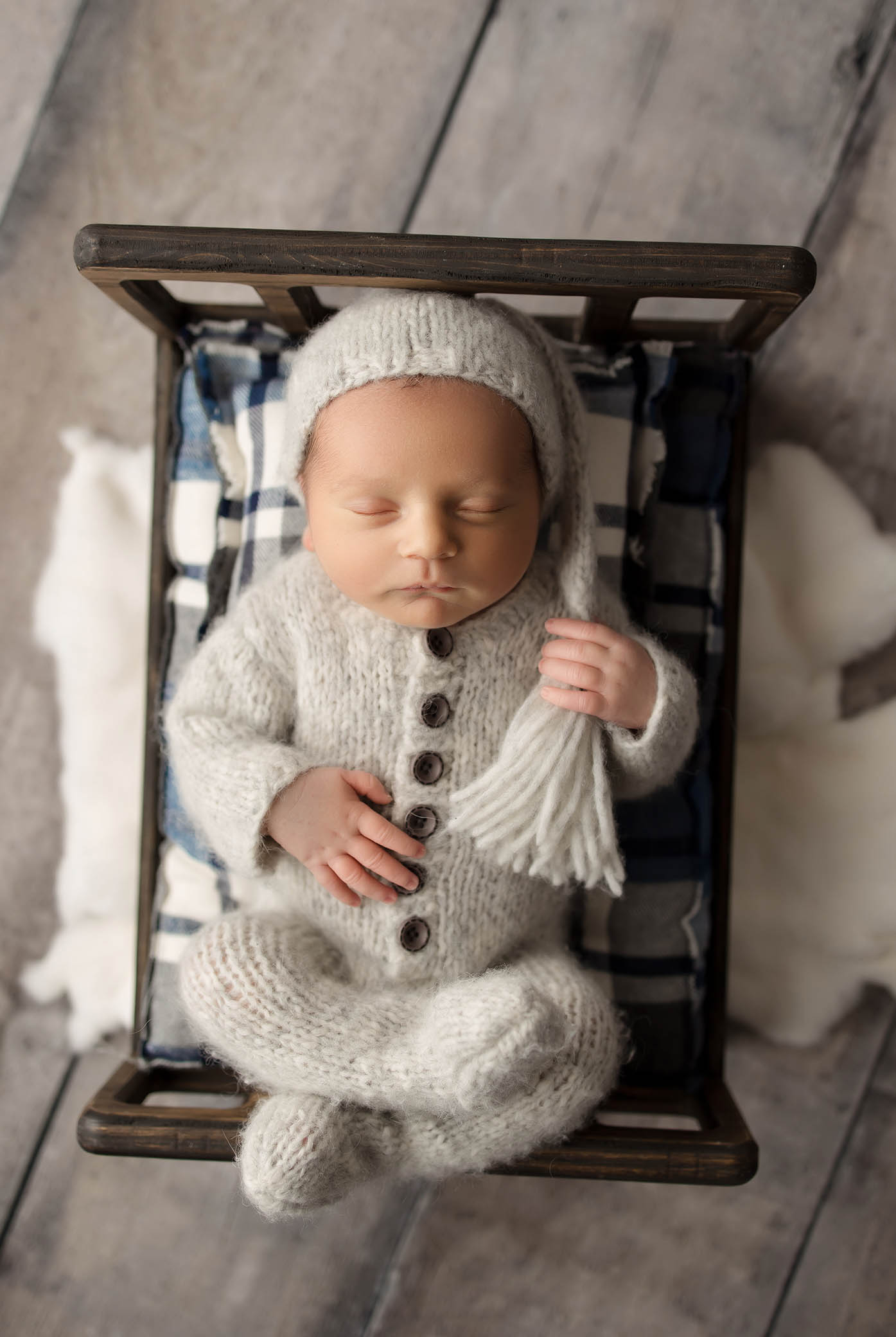 Sleeping newborn in white knitted outfit and hat on wooden bed with plaid blanket and cushion.
