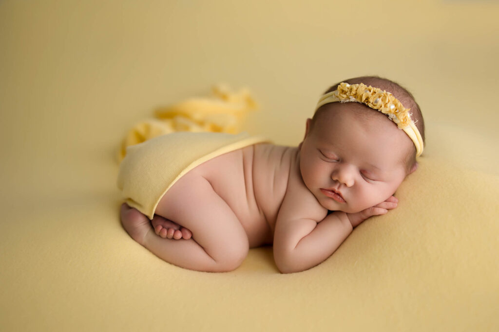 Sleeping newborn on a yellow blanket, dressed in yellow wrap and headband, hands under face.
