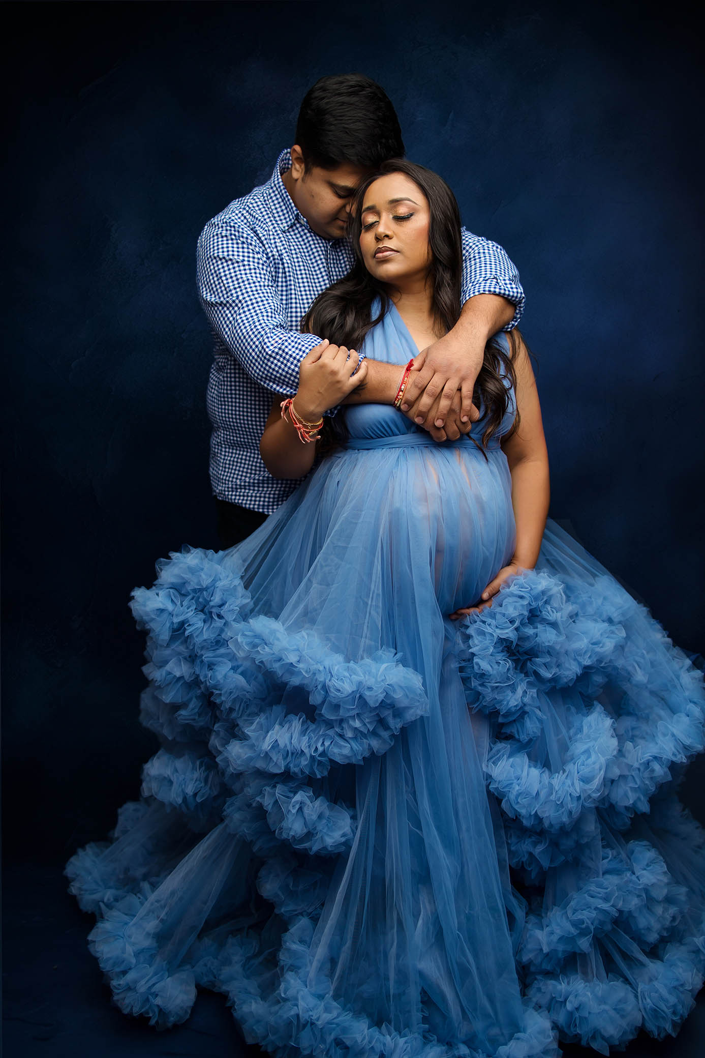 Pregnant woman in flowing blue gown embraced from behind by a man, dark blue background.