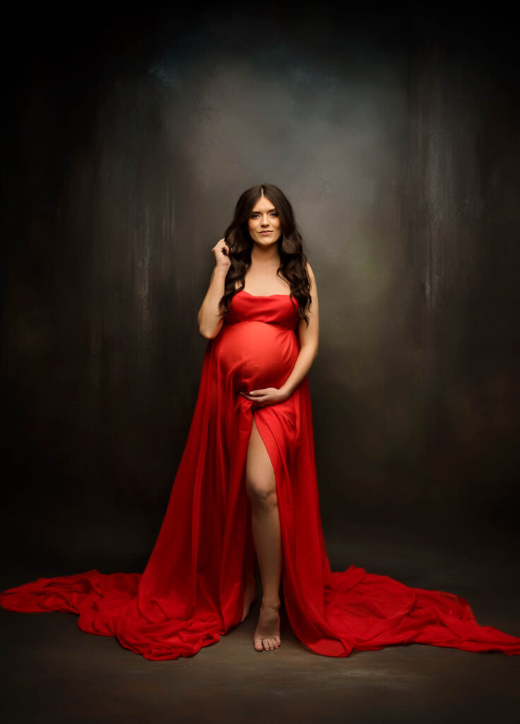 Pregnant woman in long red dress stands barefoot, one hand on belly, against dark studio backdrop.