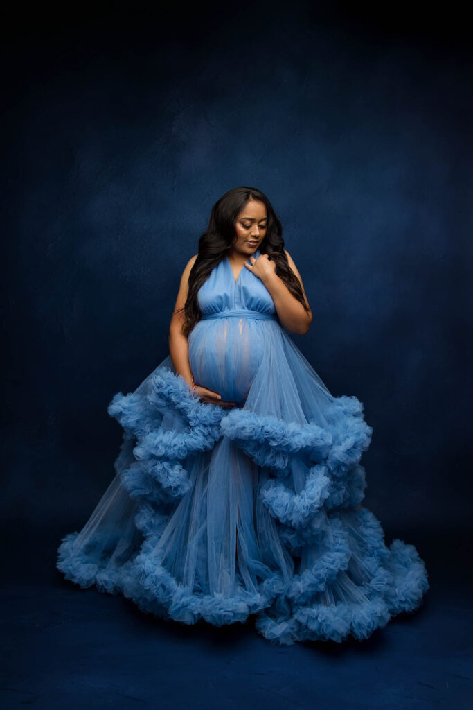Pregnant woman in a ruffled blue gown holds her belly, gazing down against a dark background.